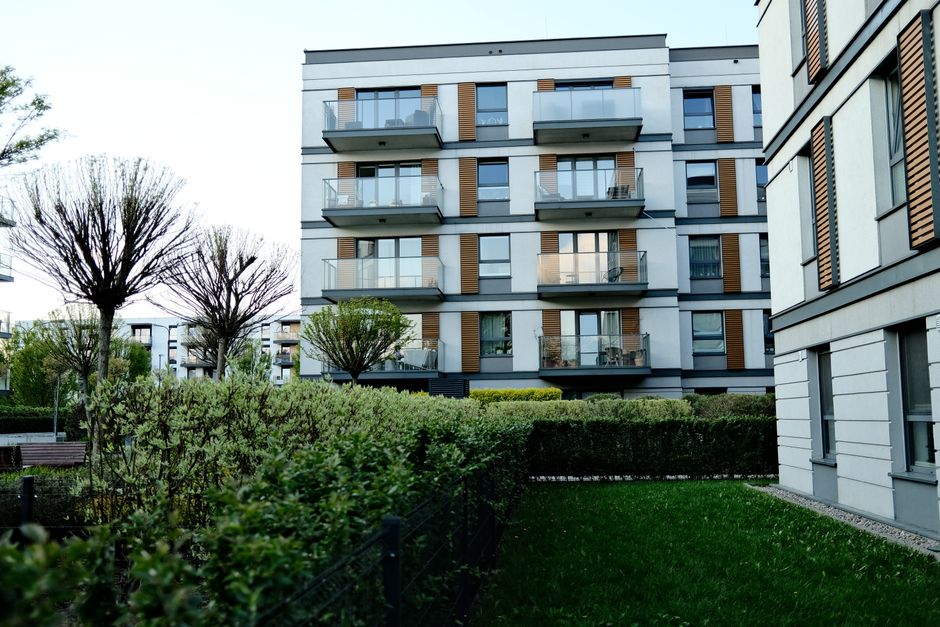 Exterior view of a modern multi-storey residential building with white facade and wooden accents, featuring glass balcony railings. Surrounding the building is a well-maintained garden with trimmed bushes, a variety of trees, and a green lawn. The scene is illuminated by natural daylight, highlighting the cleanliness and orderliness of the outdoor area. The image emphasizes the importance of outdoor surface maintenance and tidiness in residential environments, reflecting the services provided by Cleaners W12 for estate cleaning and property maintenance in Shepherds Bush Green, W12.