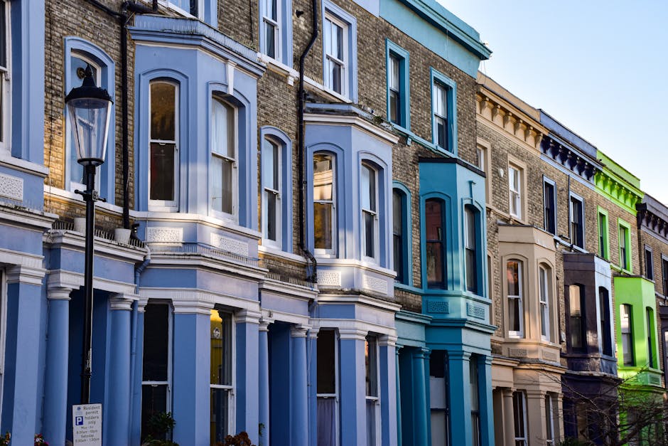 Photograph of a row of Victorian-style terraced houses featuring varying colorful facades in shades of light blue, teal, beige, dark gray, and bright green. The buildings have large, white-framed sash windows and decorative cornices, with some windows reflecting the sky and surrounding architecture. A black vintage-style street lamp stands in the foreground, and the clear sky above indicates daytime. The street appears clean and well-maintained, representing an urban residential area in Shepherds Bush Green. This image exemplifies the vibrant street scene that residents may wish to keep tidy through professional cleaning services like those offered by Cleaners W12.